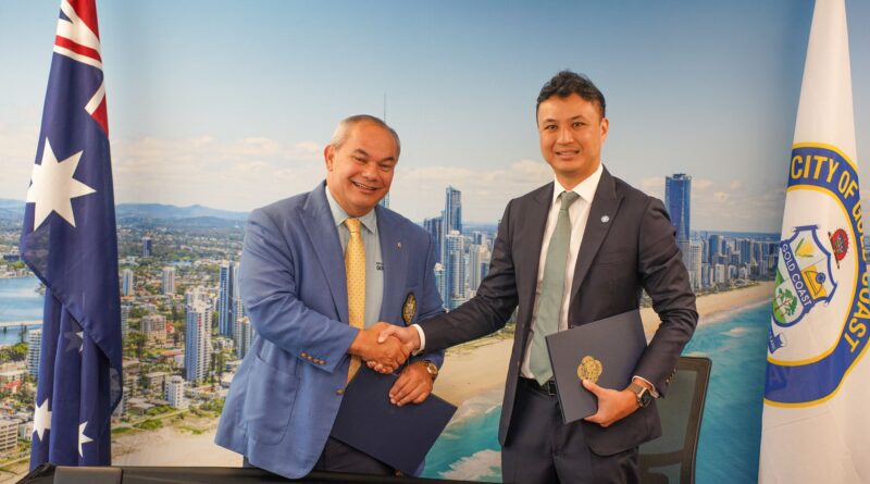 Two men in business suits shake hands in front of a city skyline backdrop, each holding a folder or document.
