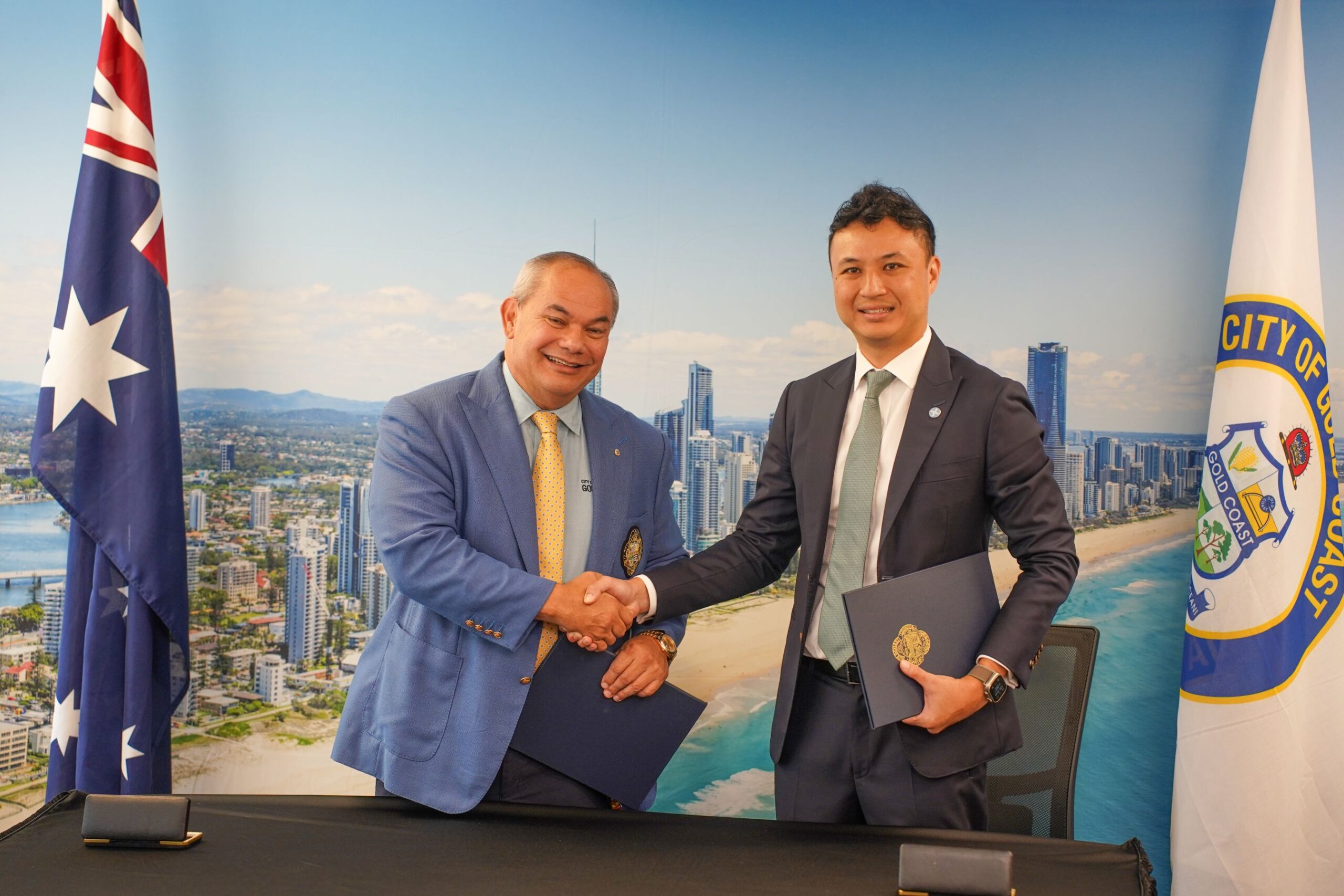 Two men in business suits shake hands in front of a city skyline backdrop, each holding a folder or document.