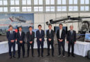 Group photo - eVTOL Insights Seven men in dark suits posing for a group photo inside an aviation hangar with a large helicopter on display behind them and a banner reading 'SoraCruise'.