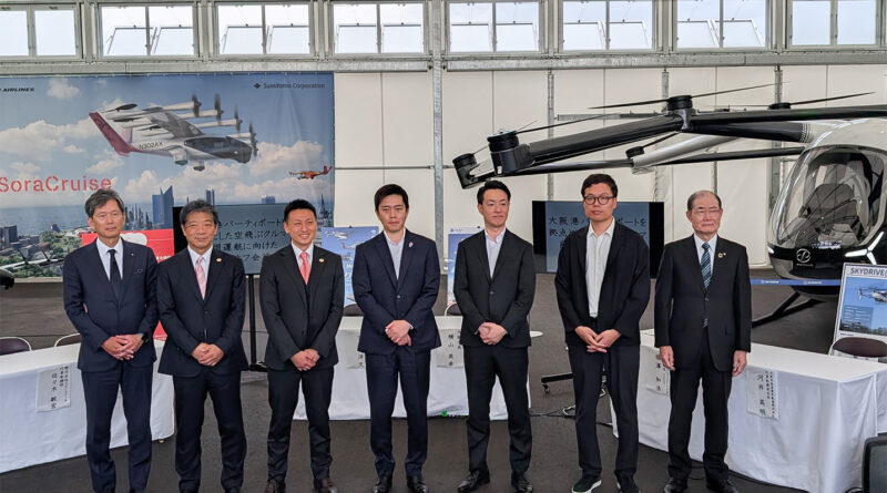 Seven men in dark suits posing for a group photo inside an aviation hangar with a large helicopter on display behind them and a banner reading 'SoraCruise'.