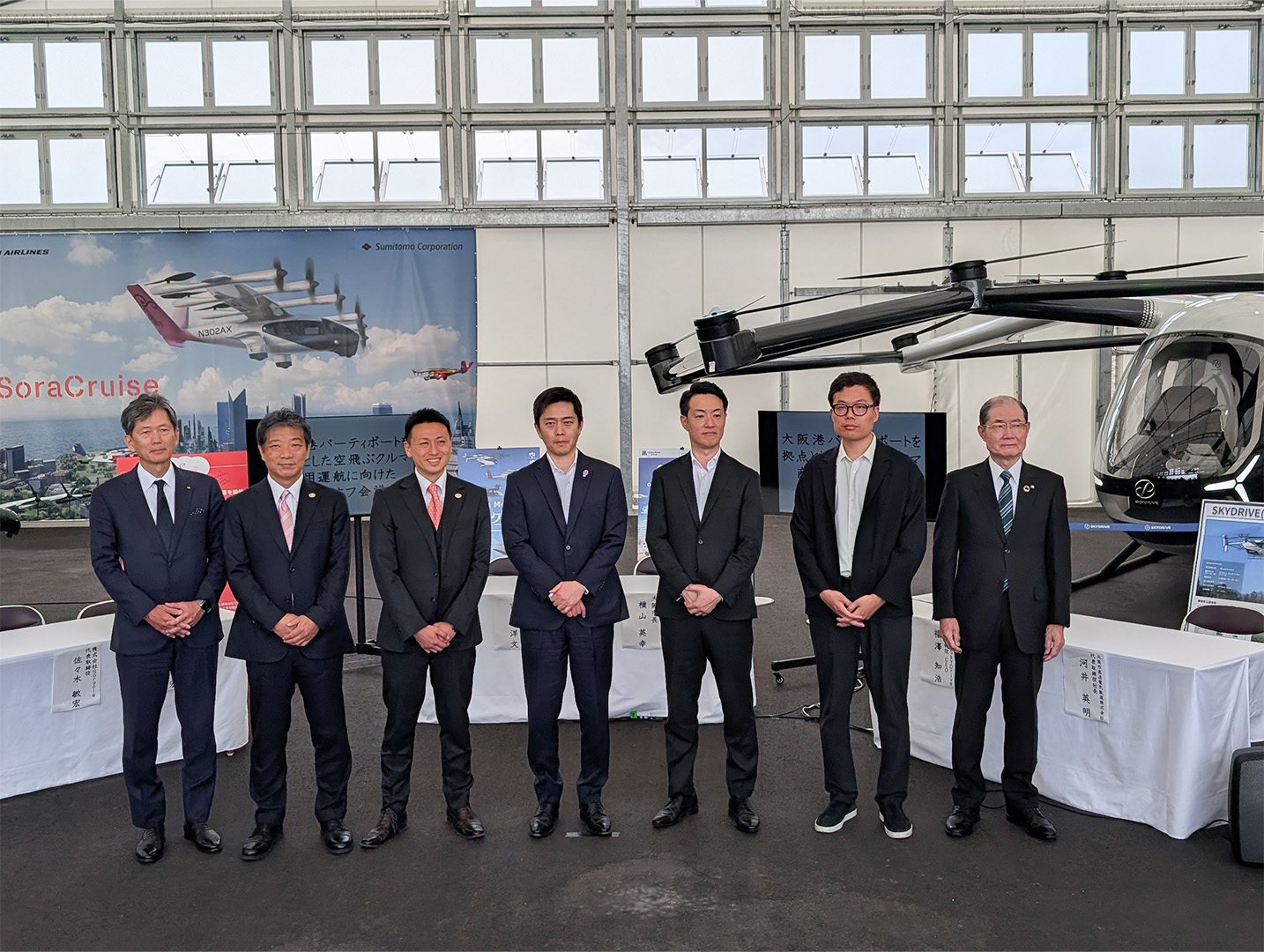 Seven men in dark suits posing for a group photo inside an aviation hangar with a large helicopter on display behind them and a banner reading 'SoraCruise'.