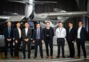 Hyundai WIA - eVTOL Insights Group of eight professionals in suits posing in front of a silver helicopter inside a hangar.