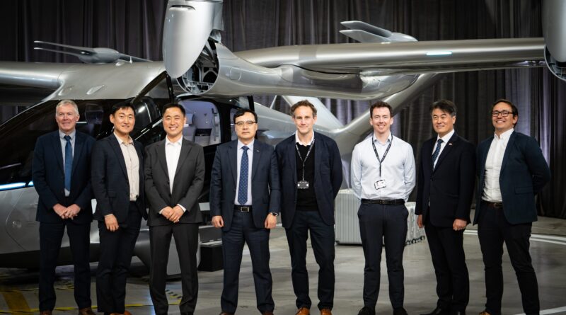 Hyundai WIA - eVTOL Insights Group of eight professionals in suits posing in front of a silver helicopter inside a hangar.