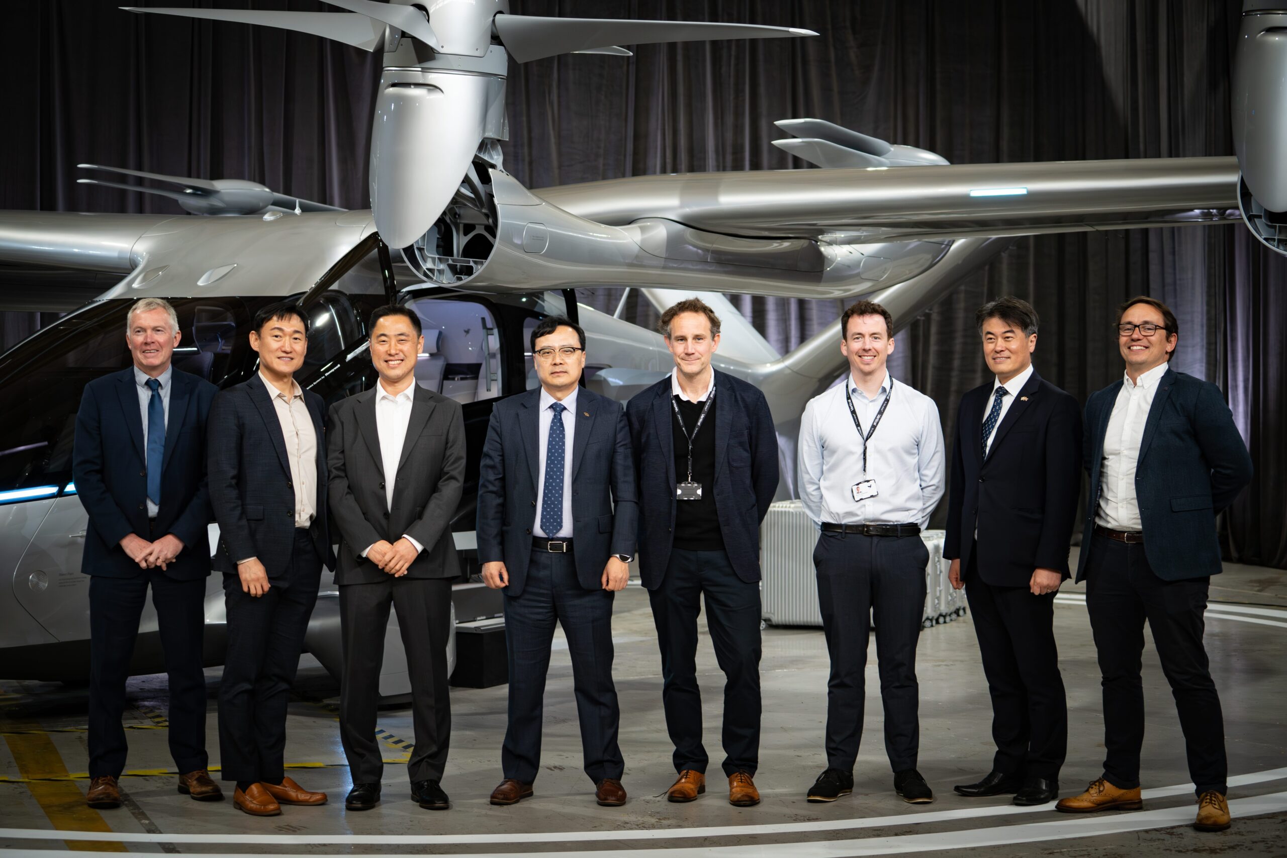 Group of eight professionals in suits posing in front of a silver helicopter inside a hangar.