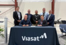 Seven men in business attire pose behind a signing table with a ViaSat banner at an aerospace event.