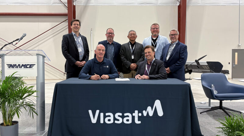 Seven men in business attire pose behind a signing table with a ViaSat banner at an aerospace event.