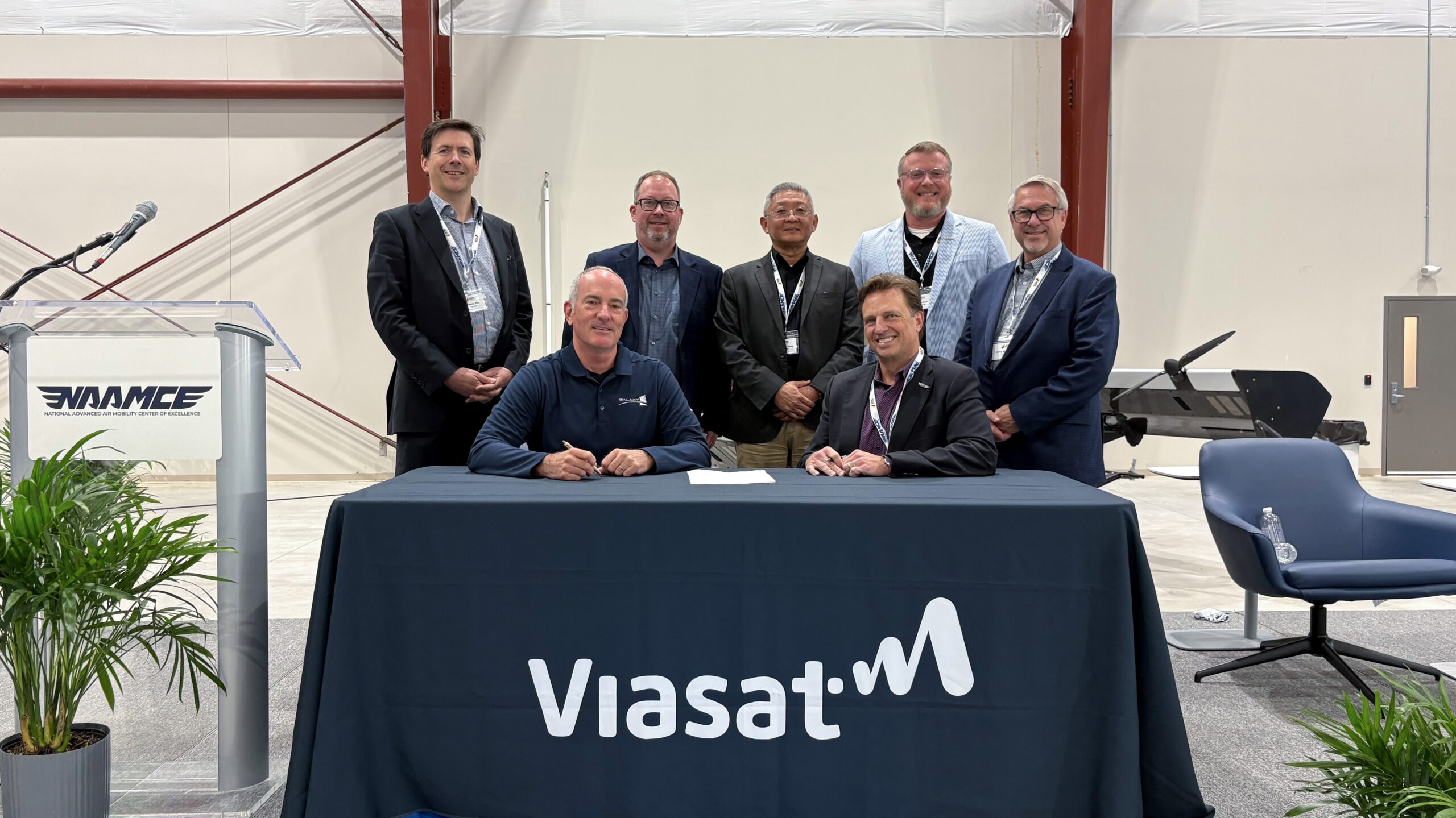 Seven men in business attire pose behind a signing table with a ViaSat banner at an aerospace event.