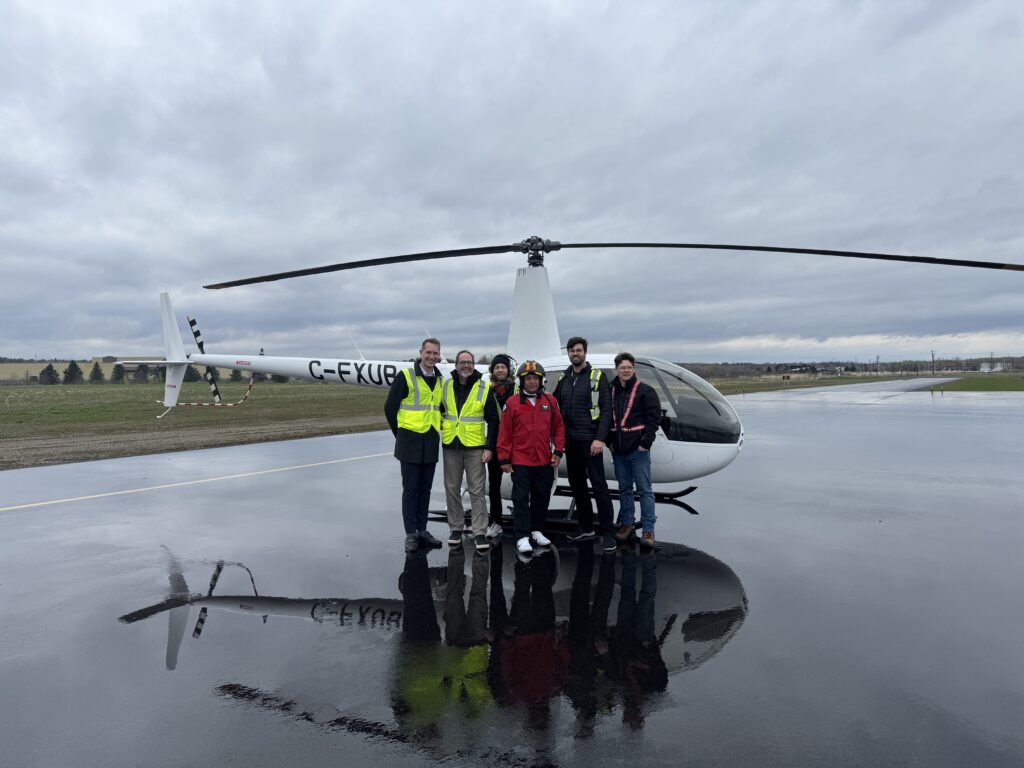 Group of people in high-visibility vests posing next to a white helicopter on a wet tarmac; reflection visible in a puddle, cloudy sky above.
