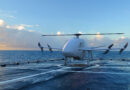 White quadcopter drone on a windswept deck with the ocean and cloudy sky in the background.