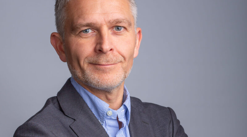 Portrait of a mature man in a gray suit with a blue striped shirt, arms crossed, neutral gray background.
