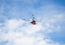 White quadcopter drone with a red cargo box beneath, flying against a blue, partly cloudy sky