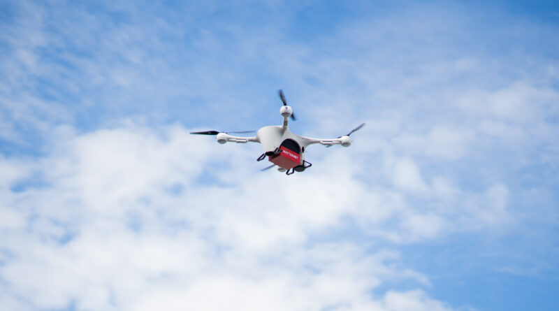 Matternet FINAL image - eVTOL Insights White quadcopter drone with a red cargo box beneath, flying against a blue, partly cloudy sky