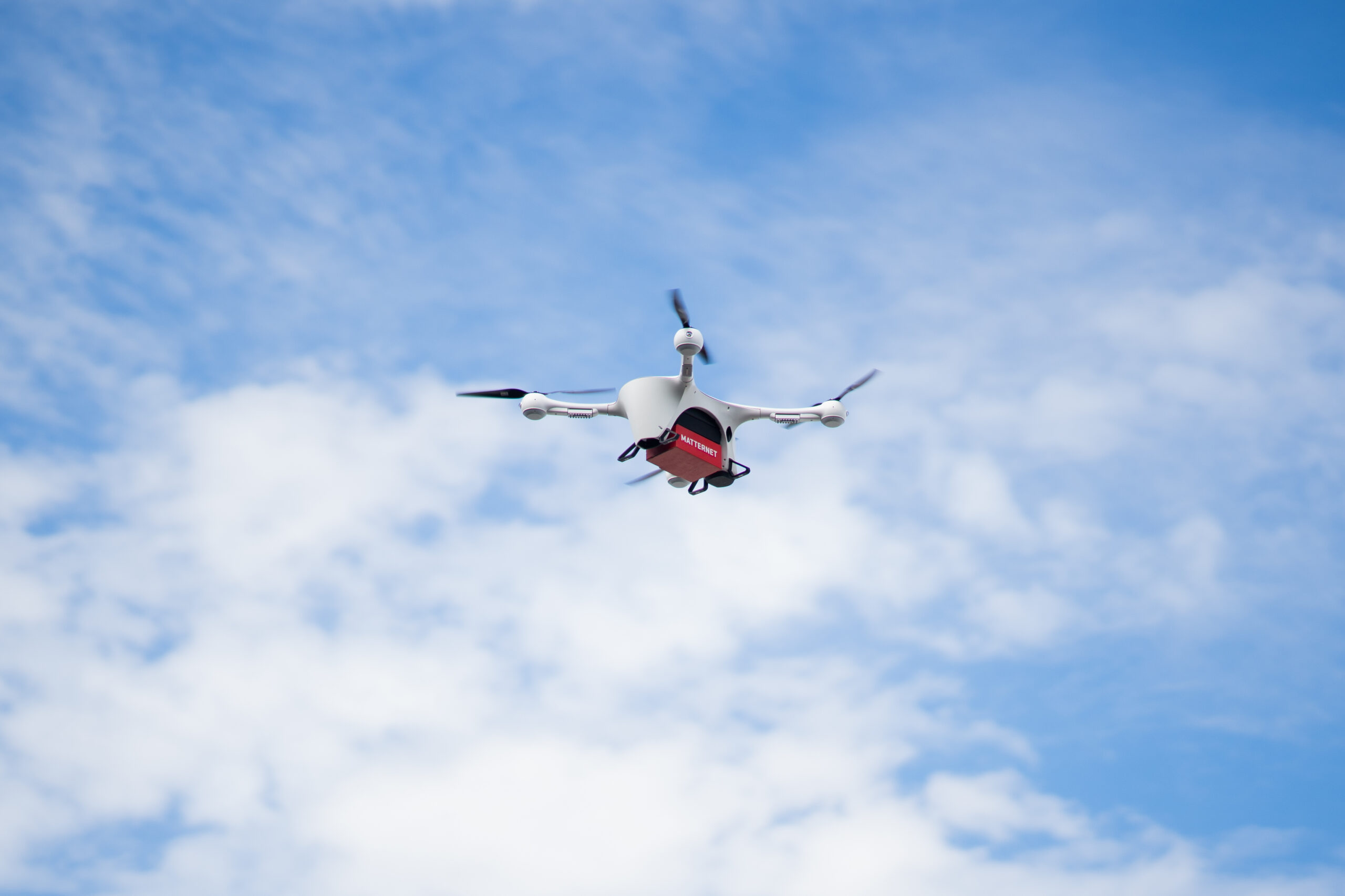 White quadcopter drone with a red cargo box beneath, flying against a blue, partly cloudy sky