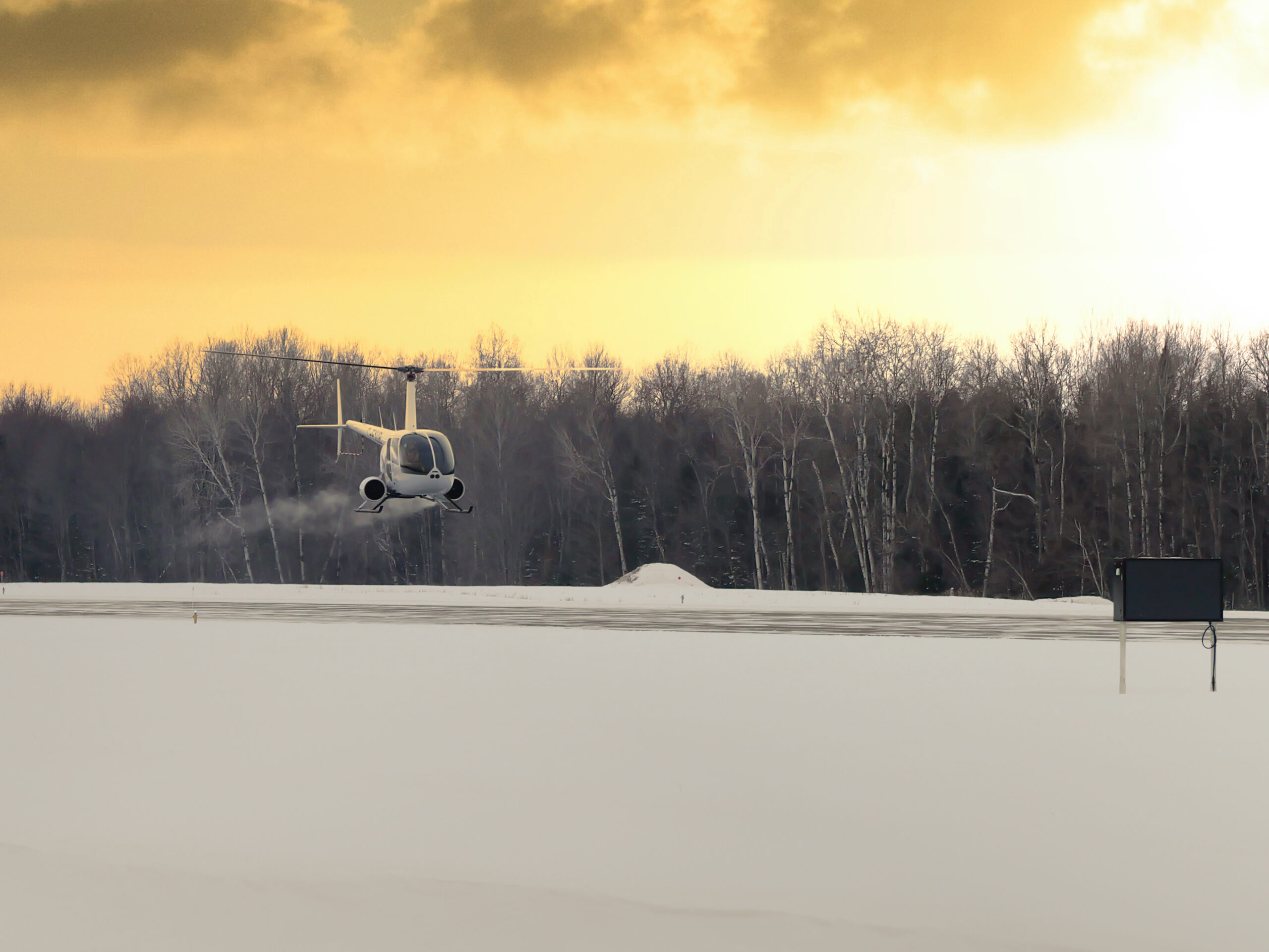 White helicopter hovering over a snow-covered field with bare trees at sunset