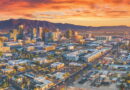 Aerial view of a downtown skyline at sunset with orange-pink clouds and distant mountains on the horizon.