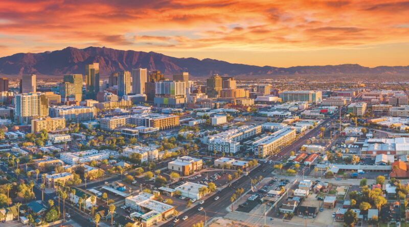 Aerial view of a downtown skyline at sunset with orange-pink clouds and distant mountains on the horizon.