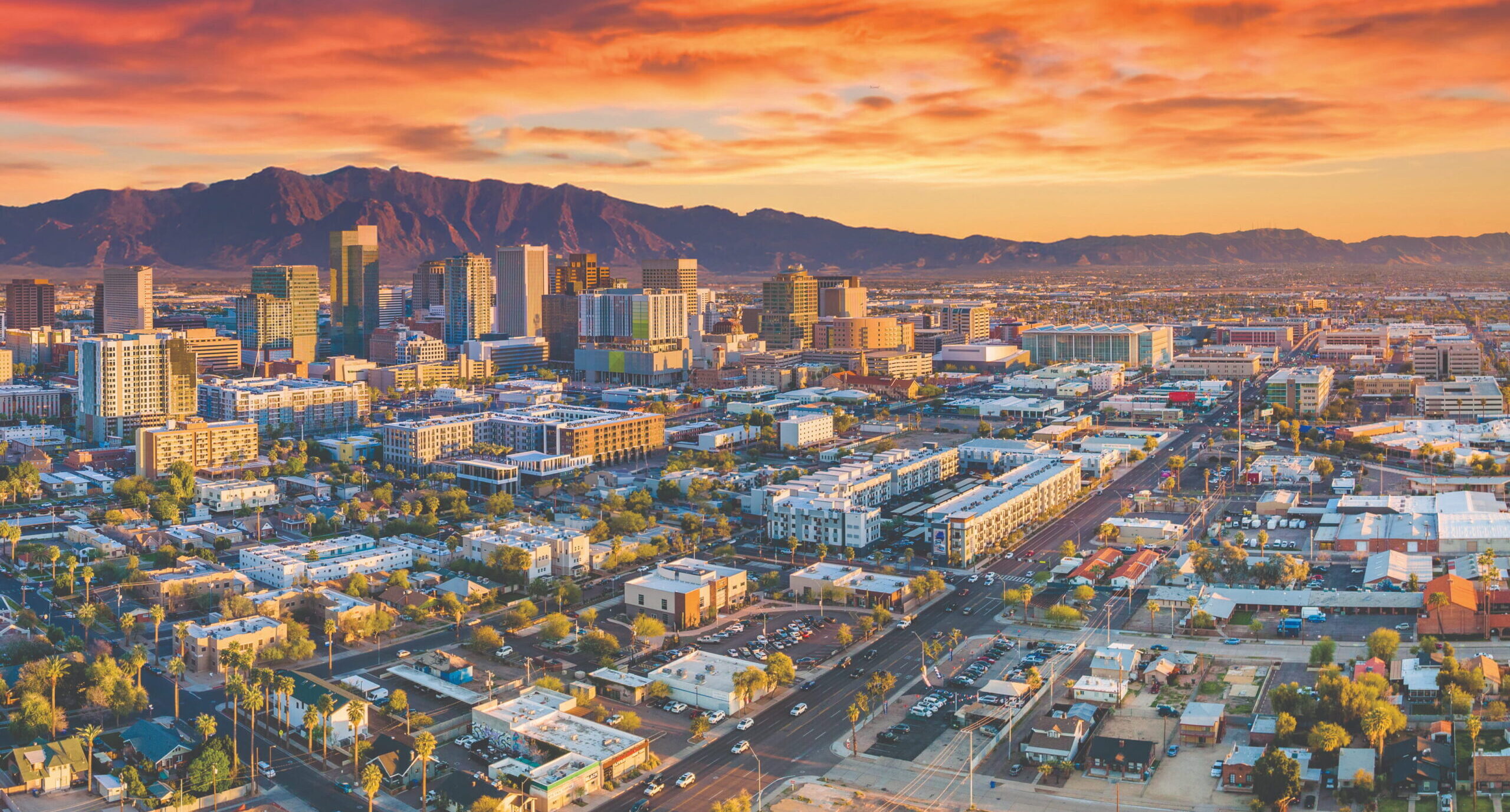 Aerial view of a downtown skyline at sunset with orange-pink clouds and distant mountains on the horizon.