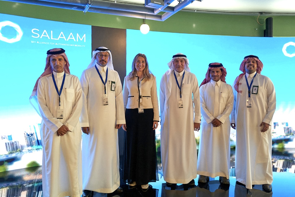 Group of six people in traditional white robes posing on a blue Salaam backdrop at an event.