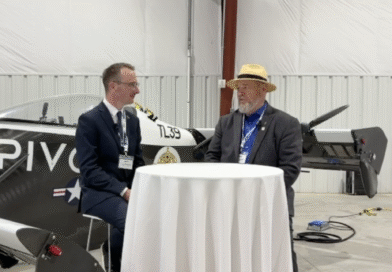 Two men in suits sit at a round table inside a hangar, with a small airplane in the background.