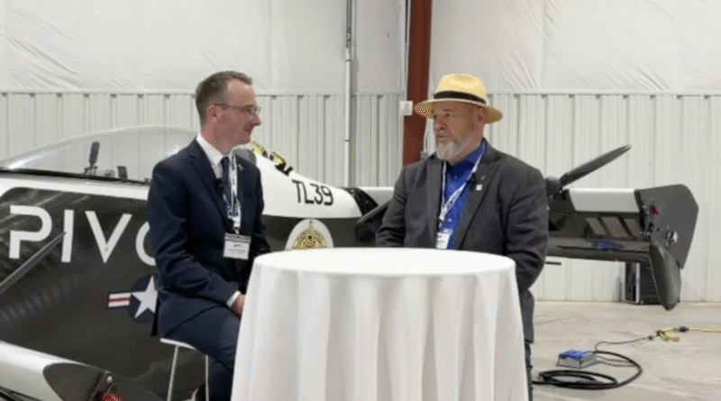 Two men in suits sit at a round table inside a hangar, with a small airplane in the background.
