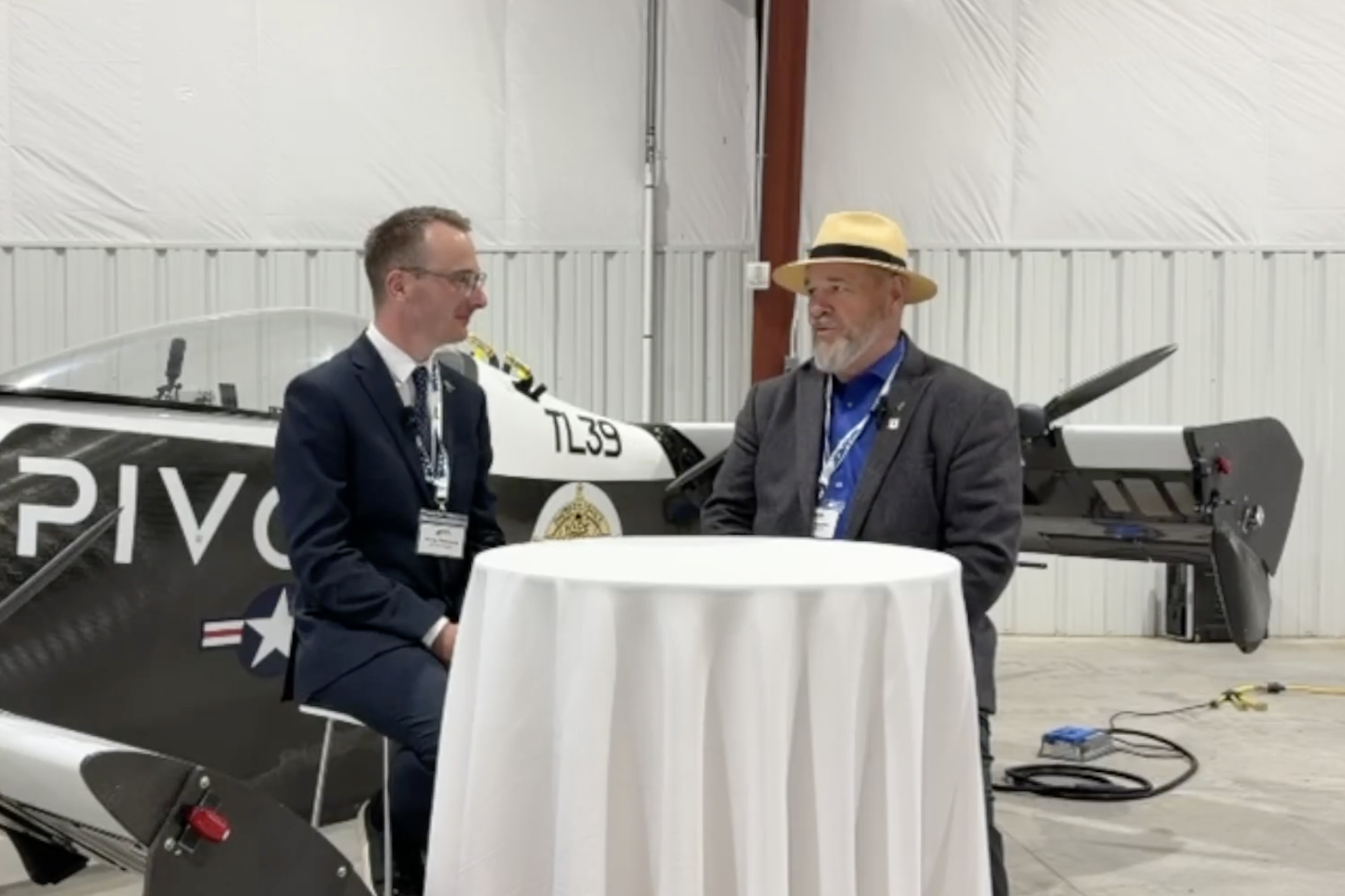 Two men in suits sit at a round table inside a hangar, with a small airplane in the background.