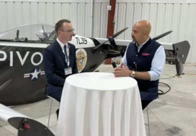Two men interview each other at a round table inside a hangar, with a propeller airplane in the background.