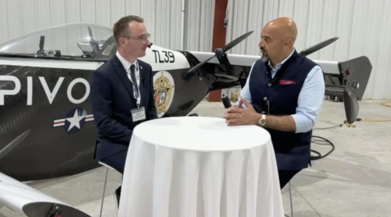 Two men interview each other at a round table inside a hangar, with a propeller airplane in the background.