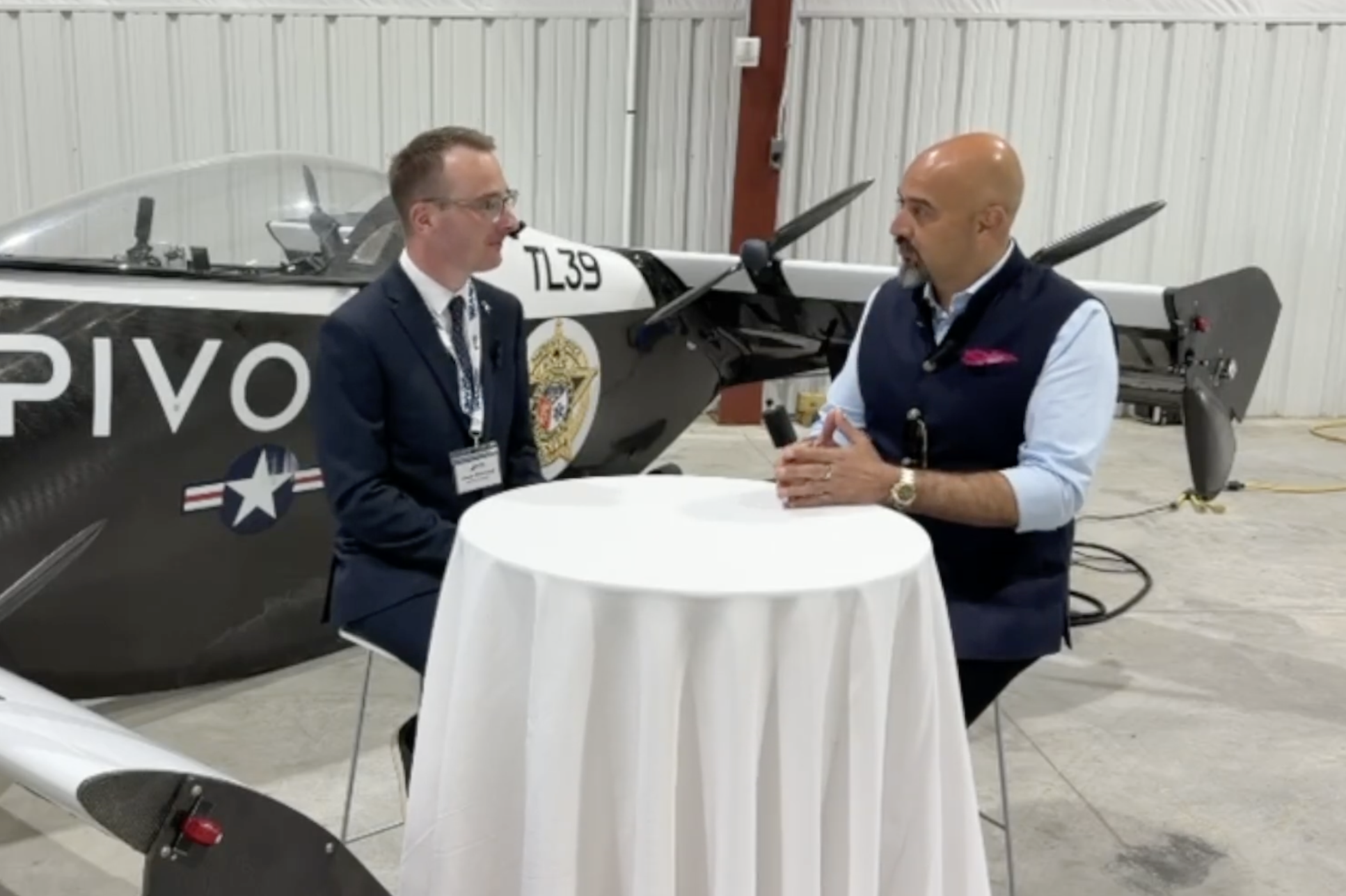 Two men interview each other at a round table inside a hangar, with a propeller airplane in the background.
