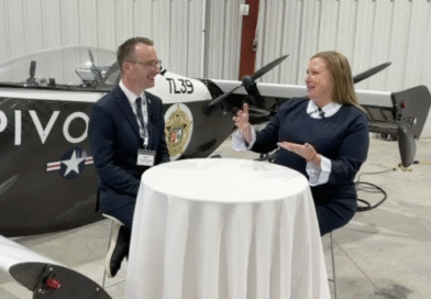 Two people in a hangar sit at a round table and talk, with a propeller airplane in the background.