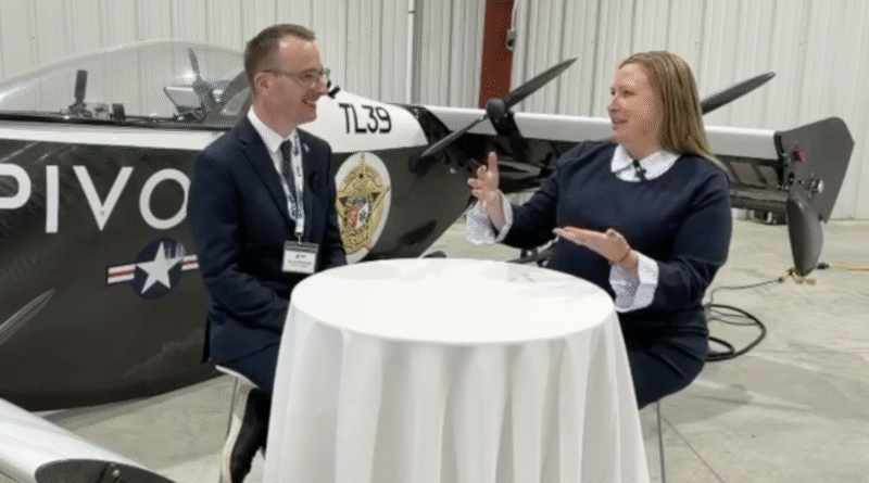 Two people in a hangar sit at a round table and talk, with a propeller airplane in the background.