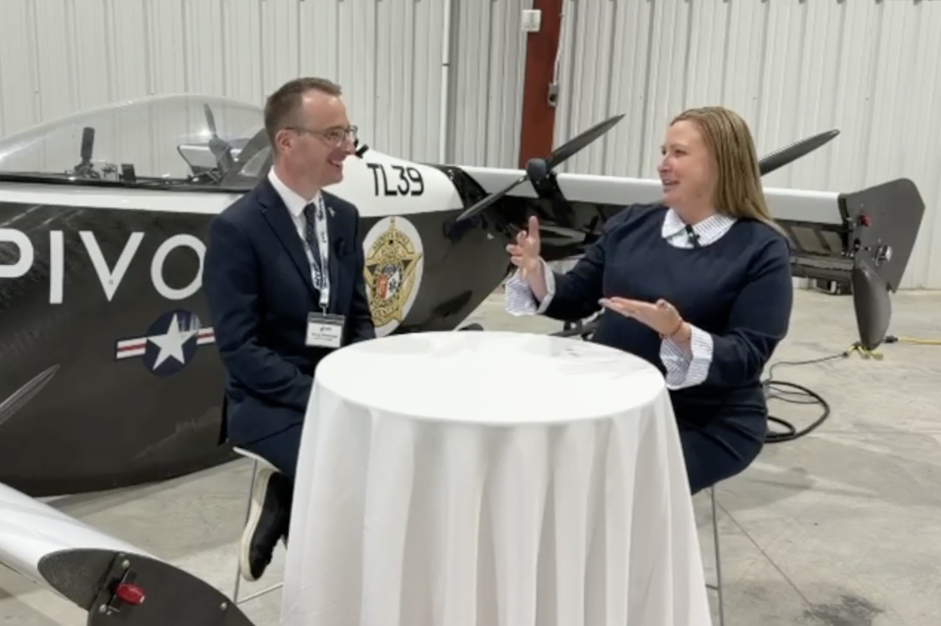 Two people in a hangar sit at a round table and talk, with a propeller airplane in the background.
