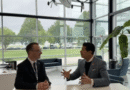 Screenshot 2026-05-18 at 163705 - eVTOL Insights Two businessmen in suits sit at a white table in a glass-walled lobby, engaged in conversation with animated hand gestures, office plant nearby.