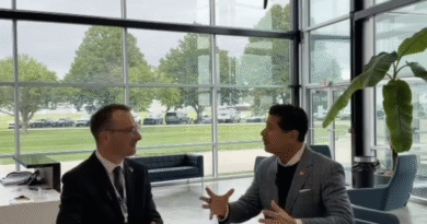 Two businessmen in suits sit at a white table in a glass-walled lobby, engaged in conversation with animated hand gestures, office plant nearby.