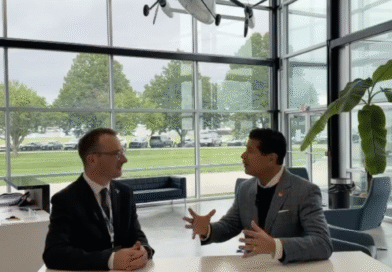 Two businessmen in suits sit at a white table in a glass-walled lobby, engaged in conversation with animated hand gestures, office plant nearby.