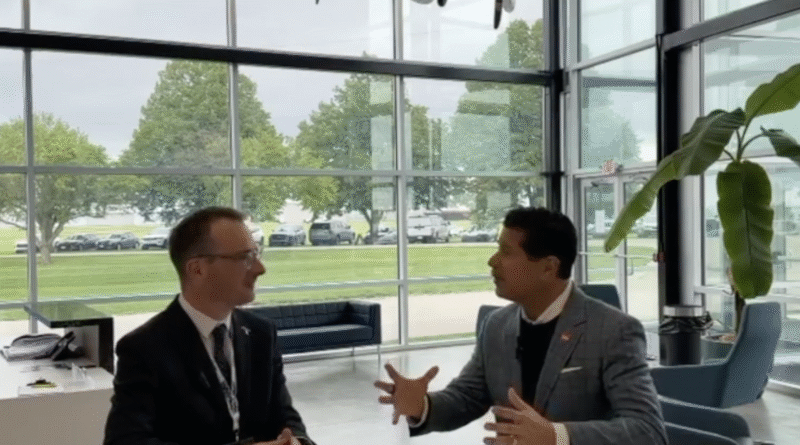 Two businessmen in suits sit at a white table in a glass-walled lobby, engaged in conversation with animated hand gestures, office plant nearby.
