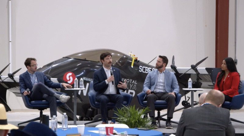 Panel discussion in an aircraft hangar with four speakers seated and a small airplane behind them.