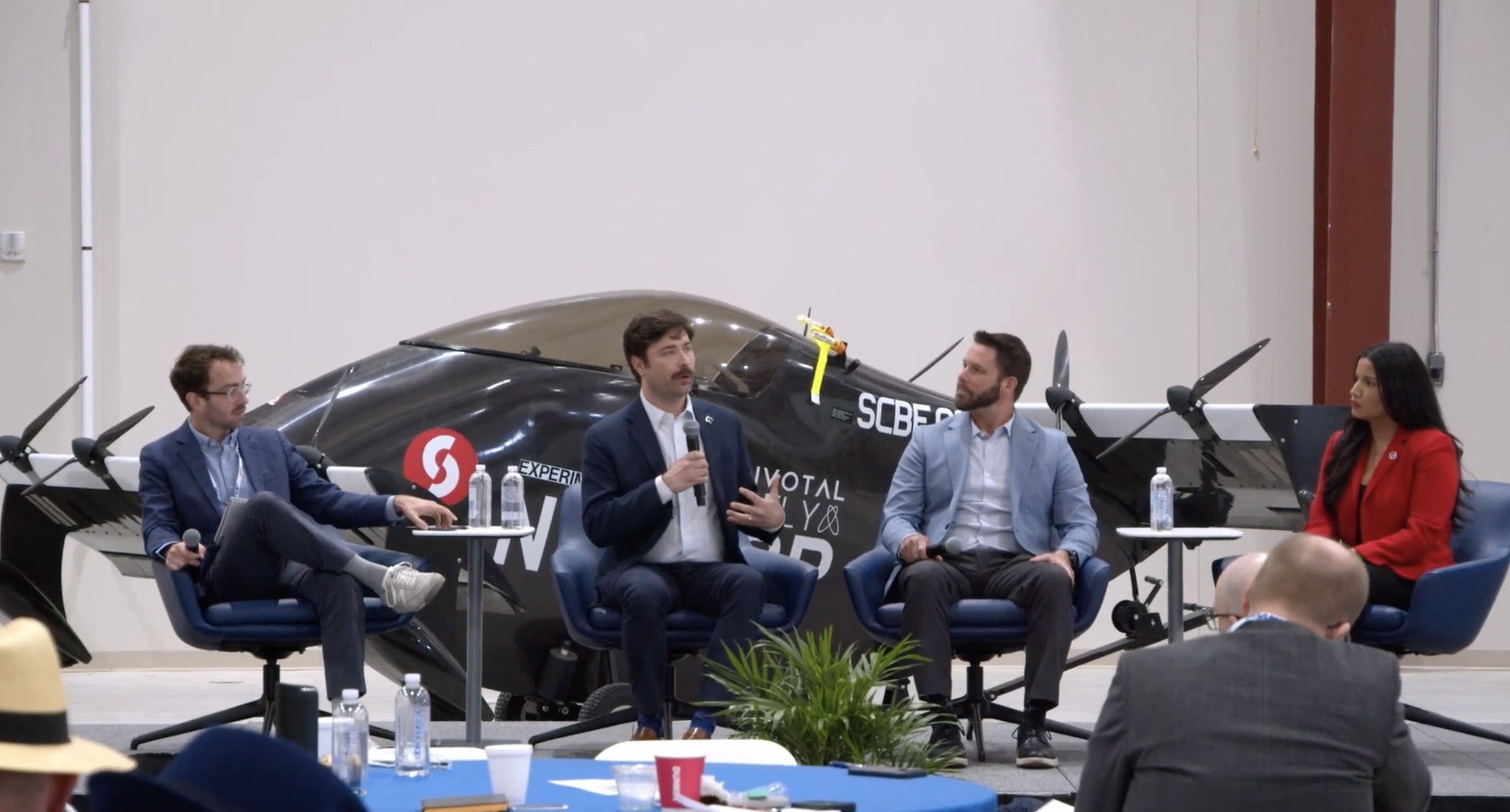 Panel discussion in an aircraft hangar with four speakers seated and a small airplane behind them.