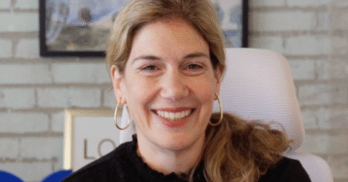 Smiling woman with blonde hair and gold hoop earrings in a black sweater, in an office setting.