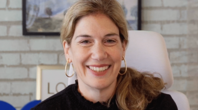 Smiling woman with blonde hair and gold hoop earrings in a black sweater, in an office setting.