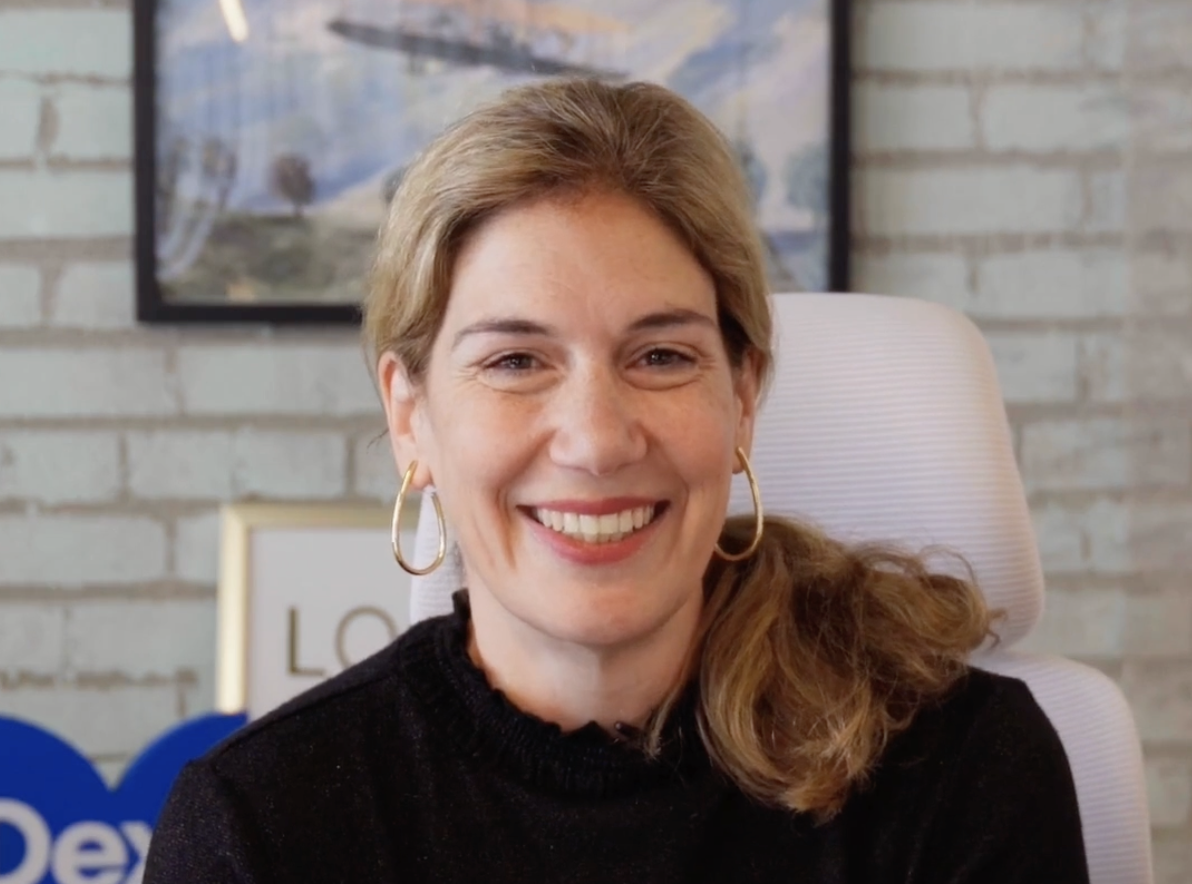 Smiling woman with blonde hair and gold hoop earrings in a black sweater, in an office setting.