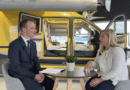 Two people sit at a round table for an interview, with a yellow helicopter and open doors behind them in a hangar, notebook and a potted plant on the table.