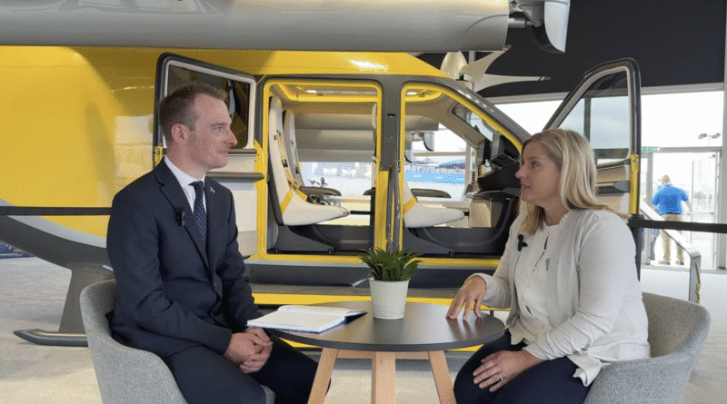 Two people sit at a round table for an interview, with a yellow helicopter and open doors behind them in a hangar, notebook and a potted plant on the table.