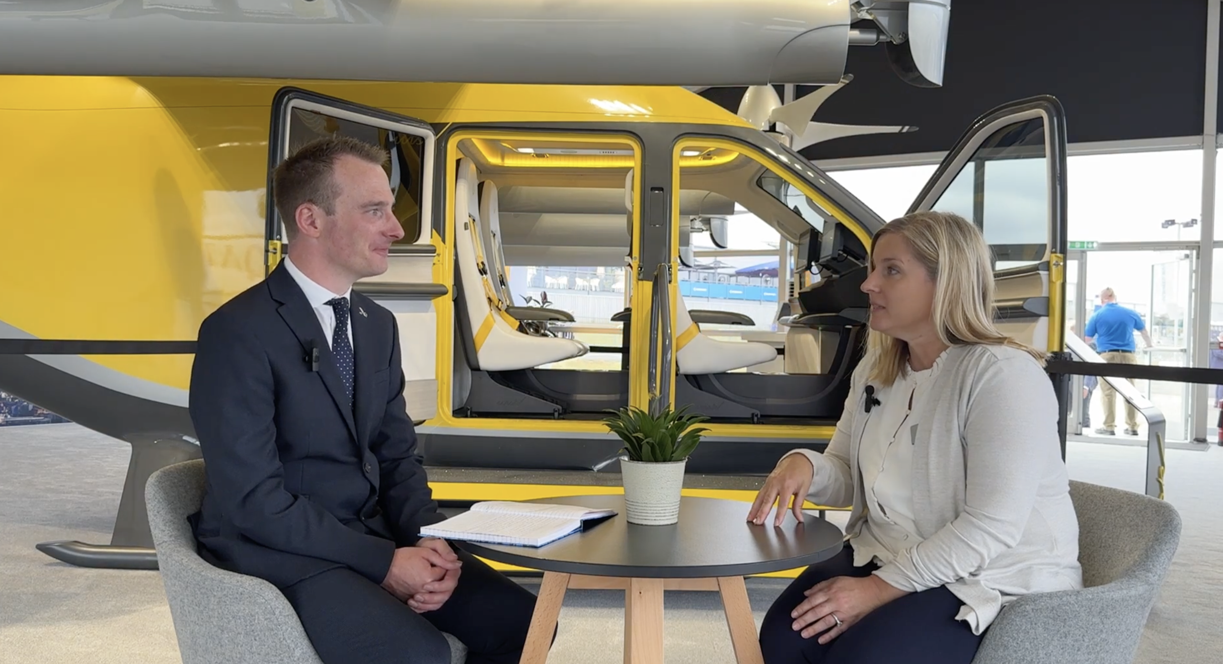 Two people sit at a round table for an interview, with a yellow helicopter and open doors behind them in a hangar, notebook and a potted plant on the table.