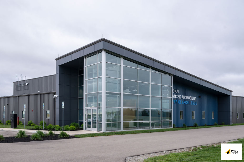 Modern industrial building with a large glass atrium and blue exterior walls; signage reads 'National Advanced Air Mobility Center of Excellence' on the side under a cloudy sky