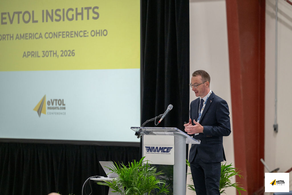 Male presenter in a navy suit speaks at a clear podium on stage during the eVTOL Insights North America Conference in Ohio (April 30, 2026).