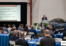 Speaker at a podium delivers a presentation to an audience; slide reads 'Ohio Aerospace and Defense Pillars' on the screen behind him.