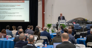 Speaker at a podium delivers a presentation to an audience; slide reads 'Ohio Aerospace and Defense Pillars' on the screen behind him.