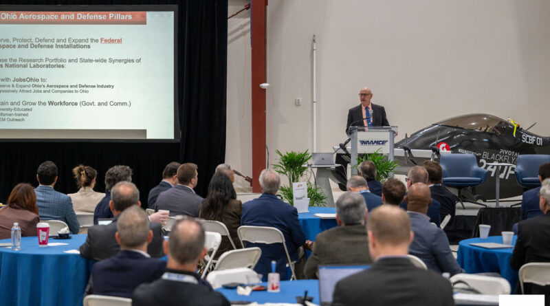 Speaker at a podium delivers a presentation to an audience; slide reads 'Ohio Aerospace and Defense Pillars' on the screen behind him.