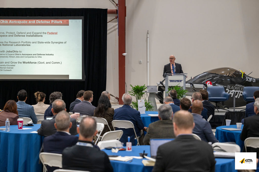Speaker at a podium delivers a presentation to an audience; slide reads 'Ohio Aerospace and Defense Pillars' on the screen behind him.