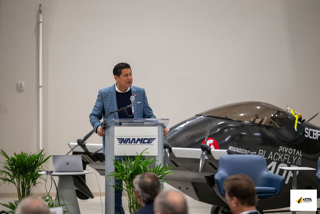 Speaker in a blue blazer at a lectern giving a talk in a hangar, with a small experimental airplane parked behind and audience members in the foreground.